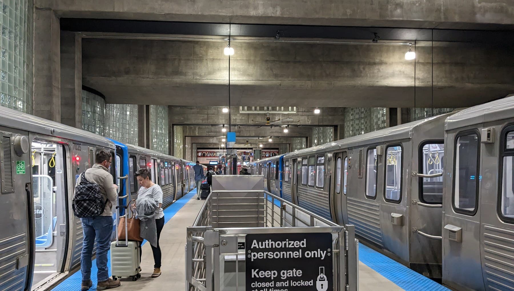 Two CTA Blue Line trains at the O'Hare terminus platform, with travellers boarding with suitcases.